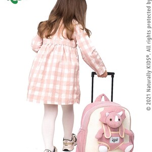 May include: A pink and cream rolling backpack with a dinosaur plushie attached. The backpack has a handle and wheels. The child is wearing a pink and white checkered dress, white tights, and sneakers. The logo "Naturally KIDS" is in the top left corner.