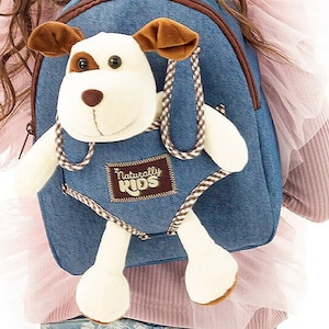 May include: A blue denim backpack with a white and brown plush dog attached to the front. The dog has brown ears and black eyes. The backpack has a brown and white checkered strap and a brown patch with the text "Naturally Kids" on it.
