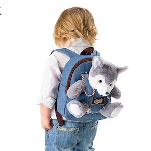May include: A young child wearing a blue denim backpack with a plush wolf attached to the front. The backpack has a brown leather strap and a brown leather tag that says "Naturally Kids".