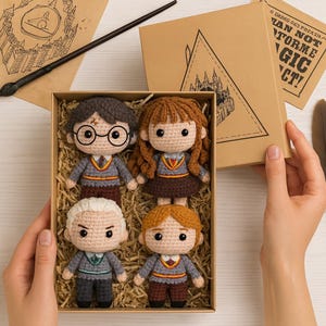 May include: A set of four crocheted dolls of characters from a popular fantasy series, presented in a brown box. The dolls have detailed features, including glasses, scarves, and uniforms. The box is filled with straw and has a lid with a triangle design.