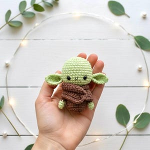May include: A small, crocheted doll in the likeness of a popular character. The doll features a light green head and large ears, with a brown cloak. The doll is held in a hand, with a white background and decorative elements.
