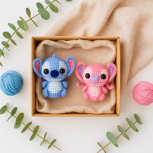 May include: Two crocheted character dolls, one blue and one pink, sit in a cardboard box lined with tan fabric. A blue yarn ball and a pink yarn ball are on either side. Green leaves surround the box.