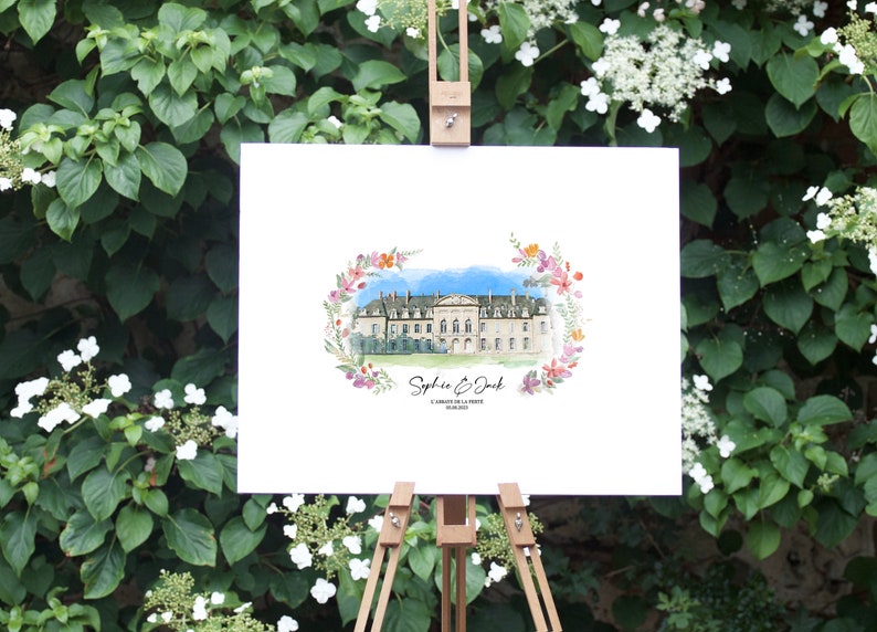 May include: A watercolor painting of a stately building, framed by a floral wreath, displayed on a wooden easel. The artwork features the names "Sophie & Jack" and a date. The background is a lush green wall of foliage.