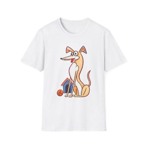 Puede incluir: Una camiseta blanca de manga corta con una ilustración de dibujos animados de un perro de color canela con hocico largo y ojos grandes, sentado junto a una caseta azul y una pelota naranja. El perro tiene el pecho blanco y una pequeña lengua roja.