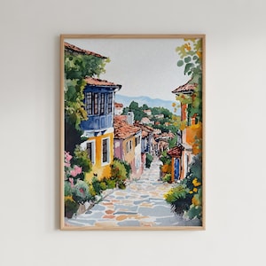 May include: Watercolor painting of a European village street scene. The artwork features colorful buildings with red tile roofs, a cobblestone street, and lush greenery. The painting is framed in a light wood frame.