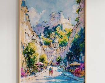 May include: Watercolor painting of a European city street scene. The artwork features a castle on a hill, buildings, trees, and people. The color palette includes blues, greens, yellows, and pinks. The painting is framed in a light-colored wooden frame.