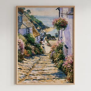 May include: A framed watercolor painting depicting a steep, stone-paved path leading down toward a coastal village. The scene features white cottages with dark roofs, lush green hillsides, vibrant pink and yellow floral bushes, and a distant sea view.