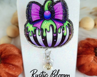 May include: A Halloween-themed badge reel in the shape of a pumpkin with a purple bow and green slime dripping down. The pumpkin has black and white stripes and small purple hearts. The text "Rustic Bloom Designs" is at the bottom.