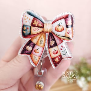 May include: A fall-themed badge reel in the shape of a bow. The bow features a patchwork design with pumpkins, leaves, and floral accents in shades of orange, red, and green. The edges are outlined with glitter. The reel has a clear plastic base.