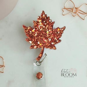 May include: A maple leaf badge reel featuring a copper-colored glitter finish. The leaf is attached to a clear plastic retractable cord and a circular base. The text "Rustic Bloom Designs" is printed in black near the bottom right corner.