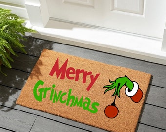 May include: A rectangular coir doormat with the words "Merry Grinchmas" in red and green. The Grinch's green hand holds a red bauble. The mat is placed in front of a white door.