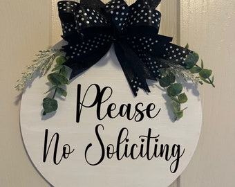May include: A round, white wooden sign with the words "Please No Soliciting" in black script. The sign is decorated with a large black bow with white polka dots, and greenery. The sign hangs from a white beaded string.