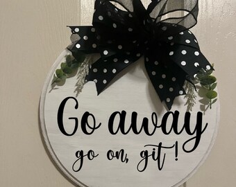 May include: A round, white wooden sign with the text "Go away go on, git!" in black script. The sign is decorated with a large black bow with white polka dots and greenery.