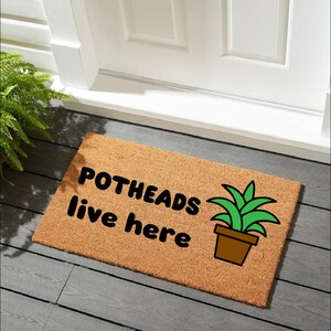 Puede incluir: Felpudo rectangular de fibra natural con el texto "POTHEADS live here" en negro y una imagen de una planta en maceta. El felpudo está colocado frente a una puerta blanca, sobre una superficie de madera gris.