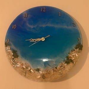May include: Round wall clock with a beach-themed design. The clock face features a gradient of blue hues, resembling the ocean, with gold-colored numerals and hands. The bottom edge is adorned with sand, shells, and small green accents.
