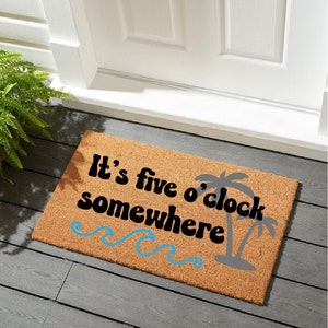 May include: A rectangular doormat made of natural fibers. The mat features the phrase "It's five o'clock somewhere" in black text, with a gray palm tree silhouette and blue wave accents. The mat is placed in front of a white door.