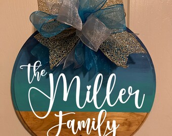 Può includere: Targa rotonda in legno con nome di famiglia con parte superiore sfumata blu e turchese e fondo in legno naturale. La targa recita "The Miller Family" in corsivo bianco, con "est. 2019" sotto. Un grande fiocco con nastri turchesi, argentati e dorati adorna la parte superiore.