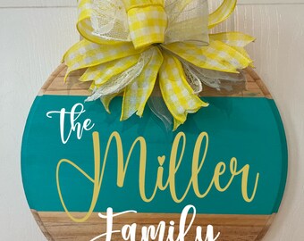 Può includere: Un cartello rotondo in legno con una sezione dipinta di turchese e la scritta "The Miller Family" in oro e bianco. Il cartello è sormontato da un grande fiocco in vichy giallo e nastro di pizzo bianco.