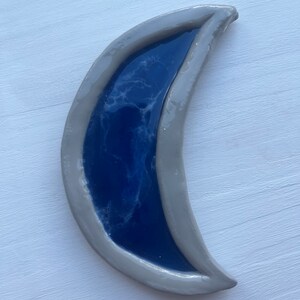 Puede incluir: Plato de cerámica en forma de media luna con borde gris e interior azul oscuro. La sección azul tiene un patrón blanco veteado. El plato está diseñado para parecerse a una media luna. El plato está sobre una superficie blanca.
