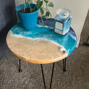 May include: Round side table with a beach-themed epoxy resin top. The table features a sandy beach design with blue and white waves, a seashell, and black hairpin legs. A blue pot with a green plant and a candle are on the table.