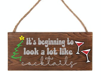 May include: A rectangular wooden sign with the text "It's beginning to look a lot like cocktails" in white and a green Christmas tree and yellow star graphic. Two red cocktail glasses are on the right. A brown rope is attached for hanging.