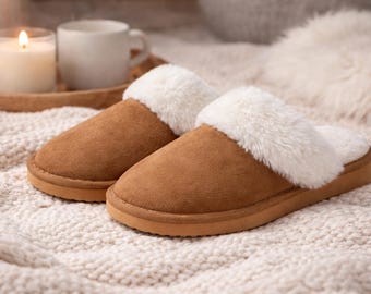 May include: A pair of brown suede slippers with a fluffy white trim. The slippers are on a textured, off-white surface, with a lit candle and a mug in the background. These cozy slippers are perfect for relaxing at home.