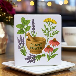 May include: White ceramic square coaster with a colorful illustration of various herbs and flowers, including lavender, mint, and yarrow, surrounding a wooden mortar and pestle. The text "PLANT POWER" is printed in green on the mortar.