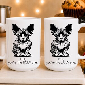 May include: Two white ceramic mugs featuring a black and white graphic illustration of a hairless Sphynx cat. Below the cat, the text reads: NO, you're the UGLY one. The mugs have a standard handle and a smooth, glossy finish on the exterior surface.