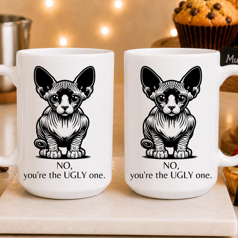 May include: Two white ceramic mugs featuring a black and white graphic illustration of a hairless Sphynx cat. Below the cat, the text reads: NO, you're the UGLY one. The mugs have a standard handle and a smooth, glossy finish on the exterior surface.