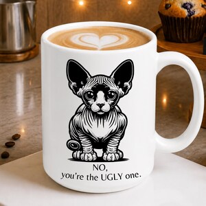 May include: A white ceramic mug filled with coffee featuring a black and white illustration of a hairless cat. Below the cat, the text reads: NO, you're the UGLY one. The mug has a large handle and is filled with a latte showing a heart-shaped foam design.