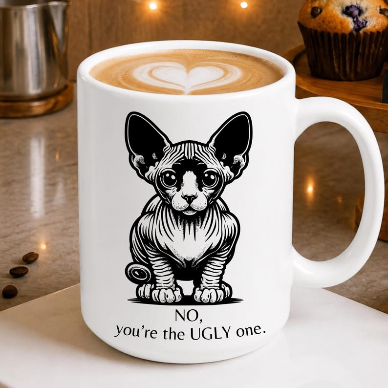 May include: A white ceramic mug filled with coffee featuring a black and white illustration of a hairless cat. Below the cat, the text reads: NO, you're the UGLY one. The mug has a large handle and is filled with a latte showing a heart-shaped foam design.