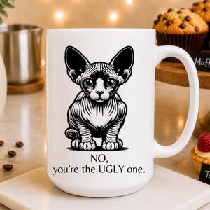 May include: A white ceramic mug featuring a black graphic illustration of a hairless Sphynx cat. Below the cat, the text reads: NO, you're the UGLY one. The mug has a standard handle and a smooth, cylindrical body design for holding beverages.