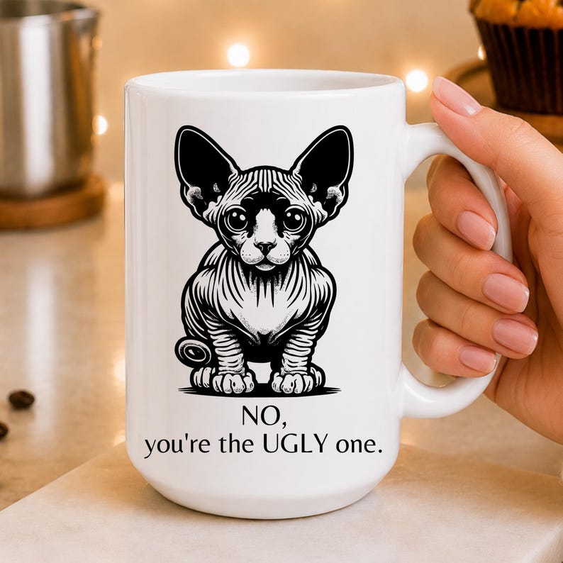 May include: A white ceramic mug featuring a black and white graphic illustration of a Sphynx cat with large ears and wrinkled skin. Below the cat, the text reads: NO, you're the UGLY one. The mug has a smooth finish and a curved handle on the right side.