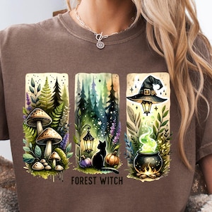May include: Brown t-shirt featuring a whimsical forest witch design. The graphic includes mushrooms, a black cat, a cauldron with green potion, and the text "FOREST WITCH". The design is in a watercolor style.