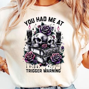 May include: A white t-shirt with a graphic design of two skulls, candles, and roses. The text "You had me at trigger warning" is printed below the graphic.