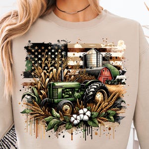 Puede incluir: Sudadera beige con un diseño de granja. El diseño incluye un tractor verde, un granero rojo y una bandera estadounidense. La imagen también presenta maíz, trigo, algodón, un silo y un molino de viento.
