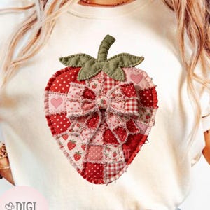May include: Cream-colored t-shirt featuring a large, patchwork strawberry design. The strawberry is composed of various red and pink fabrics, including polka dots, gingham, and heart patterns. A fabric bow adorns the center. The shirt has a round neckline.
