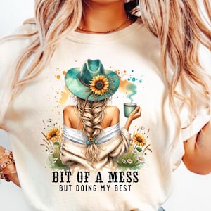 Może przedstawiać: Kremowy t-shirt z grafiką kobiety w turkusowym kapeluszu ze słonecznikiem, trzymającej kubek. Tekst "BIT OF A MESS BUT DOING MY BEST" jest wydrukowany poniżej. Na grafice znajdują się również słoneczniki i dzikie kwiaty.