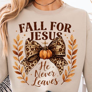 Puede incluir: Sudadera beige con el texto "FALL FOR JESUS He Never Leaves". El diseño incluye un lazo con estampado de leopardo, una calabaza, una cruz y hojas de otoño. La sudadera es de estilo cómodo e informal.