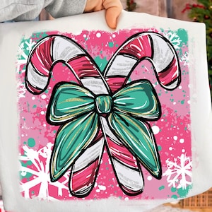 May include: A white pillow with a Christmas-themed design. The design features two candy canes with red and white stripes, a teal bow, and snowflakes on a pink background. The pillow is held by a person.