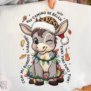Peut inclure: Sweat-shirt blanc avec un âne brodé portant un bonnet de Père Noël et un chapeau léopard. L'âne est orné de guirlandes lumineuses et entouré d'icônes festives et du texte "VAY CAMINO DE BELEN" et "CON MI BURRITO SABANERO".
