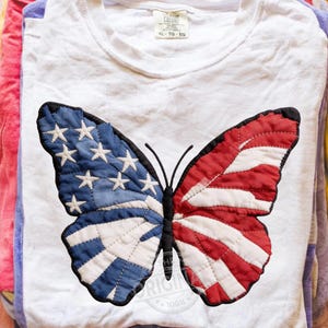 Op de afbeelding: Wit T-shirt met een vlinderontwerp. Eén vleugel toont de sterren en strepen van de Amerikaanse vlag, de andere vleugel heeft een blauwe achtergrond met witte sterren en rood-witte strepen. Het shirt heeft een label met "COLORT" en "XL - TG - EG".