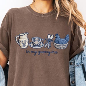 May include: Brown t-shirt featuring an embroidered design of a blue and white floral pitcher, teacup, and chicken in a basket. The text "in my granny era" is written in blue script.