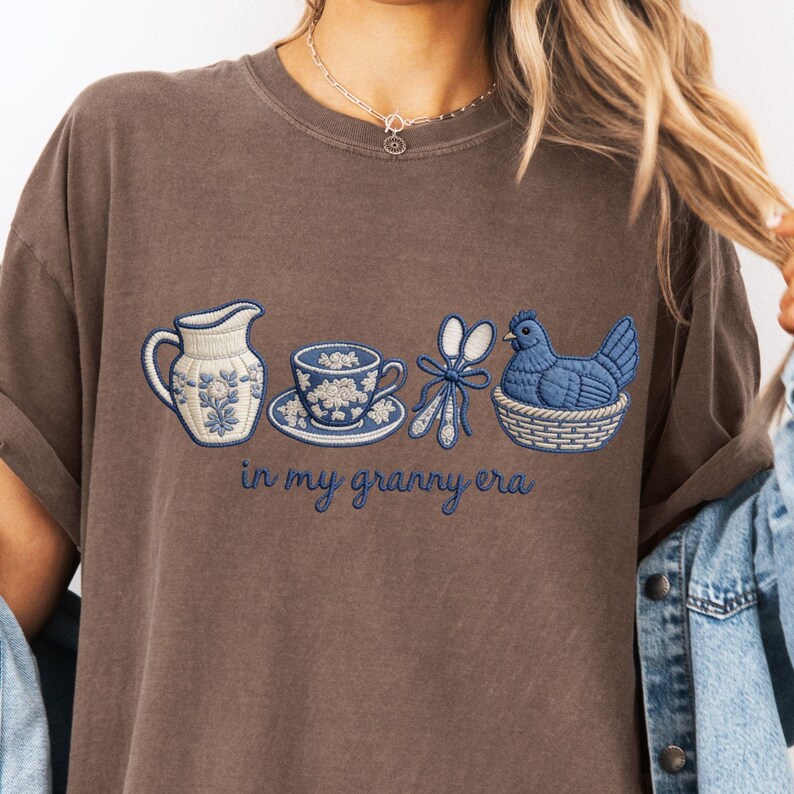 May include: Brown t-shirt featuring an embroidered design of a blue and white floral pitcher, teacup, and chicken in a basket. The text "in my granny era" is written in blue script.
