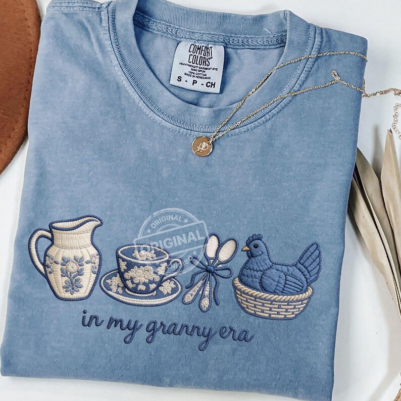 May include: A pale blue Comfort Colors t-shirt with an embroidered design of a jug, teacup, spoons, and a hen in a basket. The text "in my granny era" is below the design. A gold necklace with a pendant is also visible.
