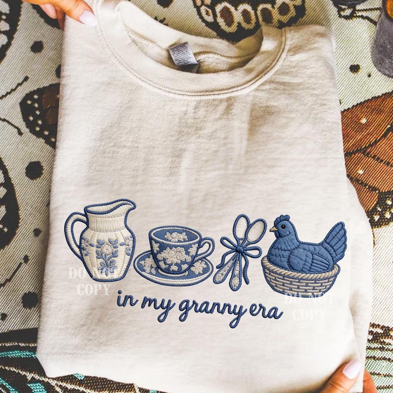 May include: A cream-coloured sweatshirt featuring blue embroidered designs: a jug, teacup, scissors, and a hen in a basket. The phrase "in my granny era" is embroidered below the designs.