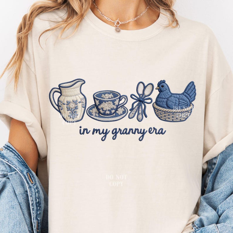 May include: A cream-coloured t-shirt with embroidered blue and white designs. The design includes a pitcher, teacup, scissors, and a hen in a basket. The words "in my granny era" are written in cursive below.