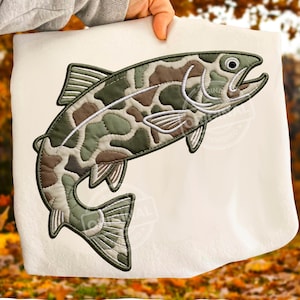 Peut inclure: Une serviette blanche avec un motif de poisson brodé. Le poisson est dans des tons de vert, marron et blanc, avec un motif de camouflage. Le poisson a des nageoires détaillées et un contour noir, créant un aspect texturé. La serviette est tenue par une personne.