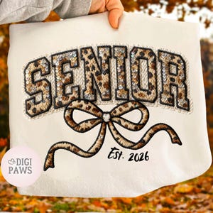 以下が含まれることがあります： ヒョウ柄のデザインで「SENIOR」の文字が施されたクリーム色のスウェットシャツ。スパンコールで装飾されています。ヒョウ柄のリボンが文字の下に刺繍され、その下に「Est. 2026」とプリントされています。スウェットシャツは人が持っています。