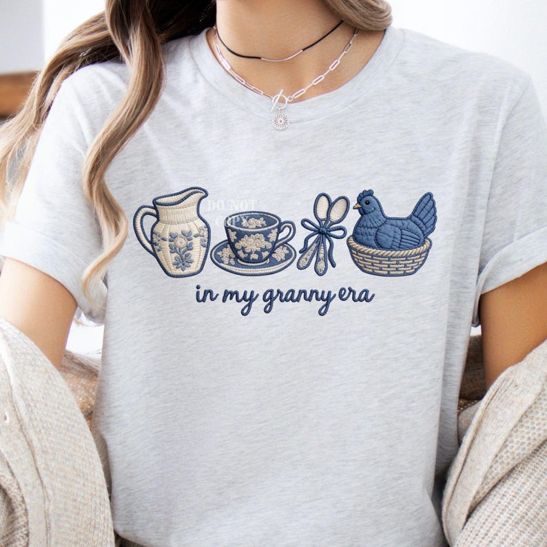 May include: Pale grey t-shirt with an embroidered design of a jug, teacup, scissors, and a hen in a basket. The text "in my granny era" is below the design. The design is in shades of blue and cream.